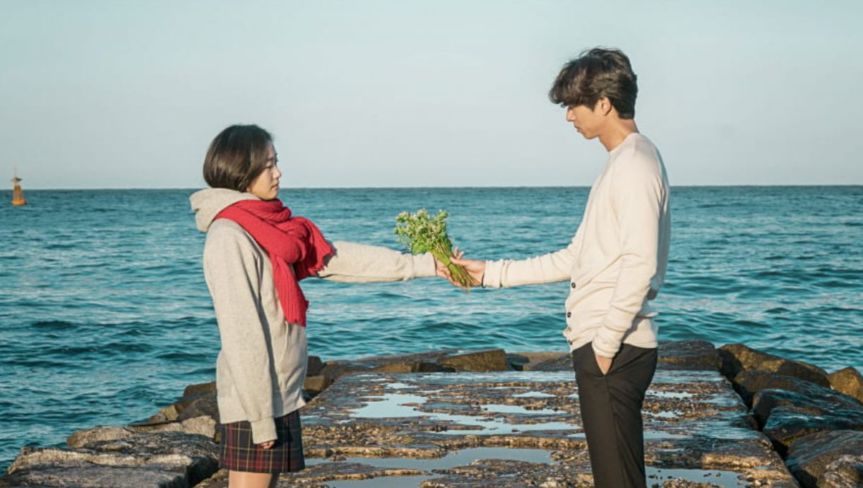 Scene from the K-drama "Goblin" where a young woman and a man stand on a stone pier by the sea, exchanging a bouquet of green flowers under a clear sky. A memorable moment highlighted on Top Honest KDrama Review Sites.