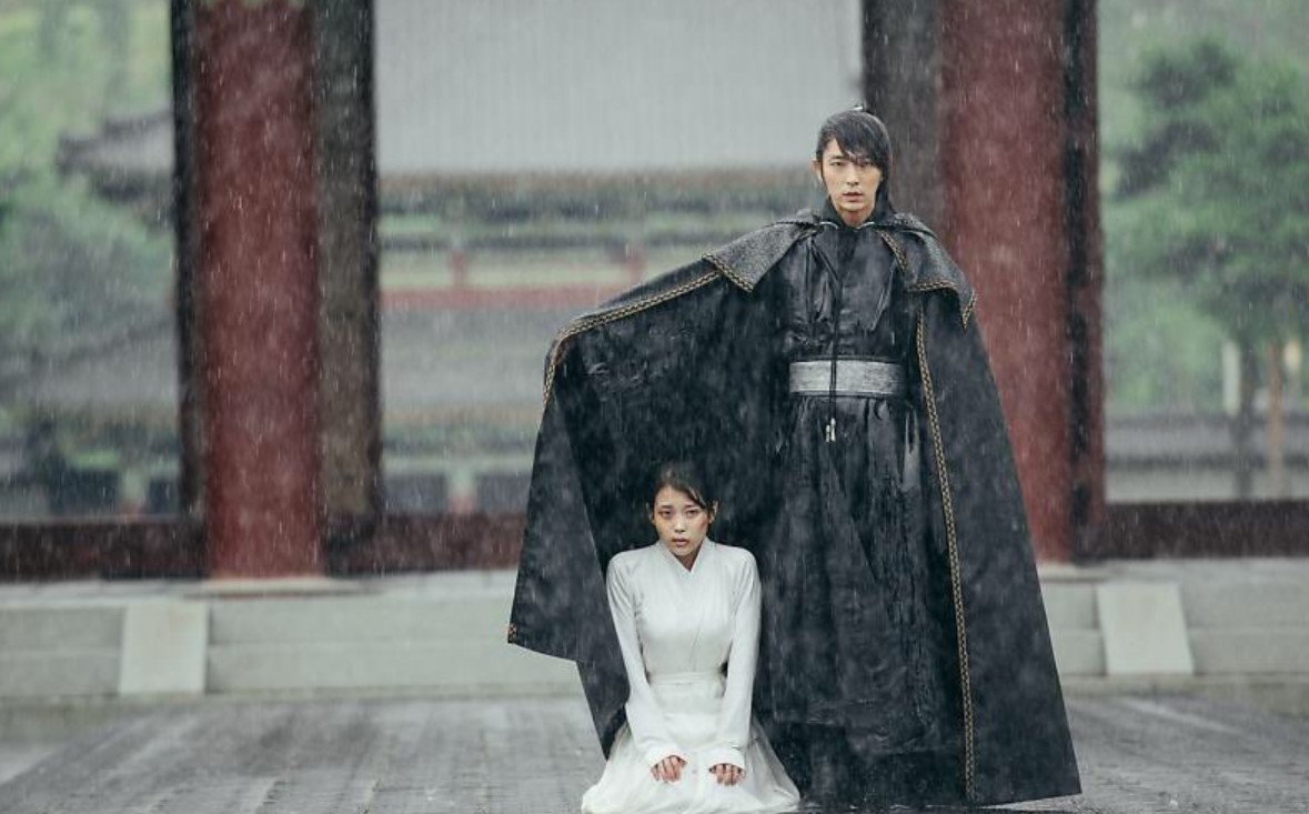 Moon Lovers: Scarlet Heart Ryeo dramatic rain scene with Lee Joon-Gi protecting IU under his cloak, as she kneels in vulnerability, highlighting the emotional intensity and protective love between the characters.