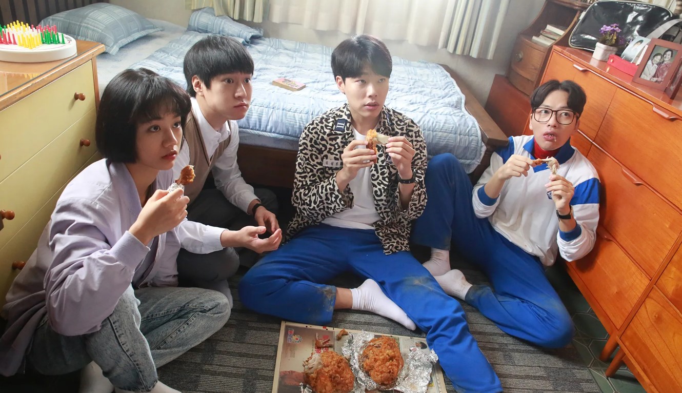 A group of friends from Reply 1988 sitting together on the floor, enjoying fried chicken and sharing a casual, fun moment in a cozy room.