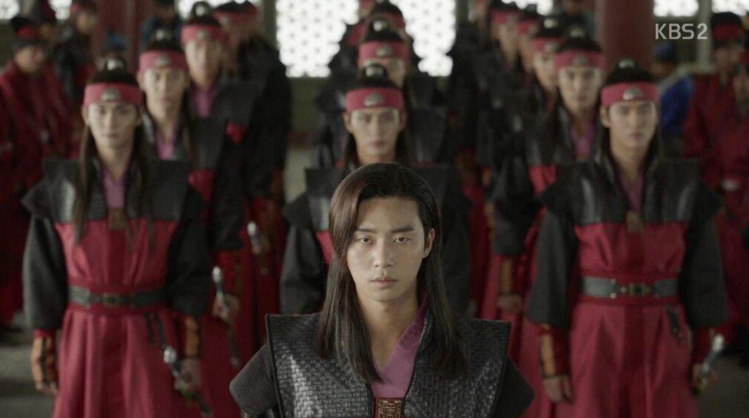 A dramatic scene from Hwarang, featuring the main character leading a group of warriors dressed in red and black uniforms. The intense focus of the characters as they stand in formation captures the serious and disciplined nature of the Hwarang warriors.