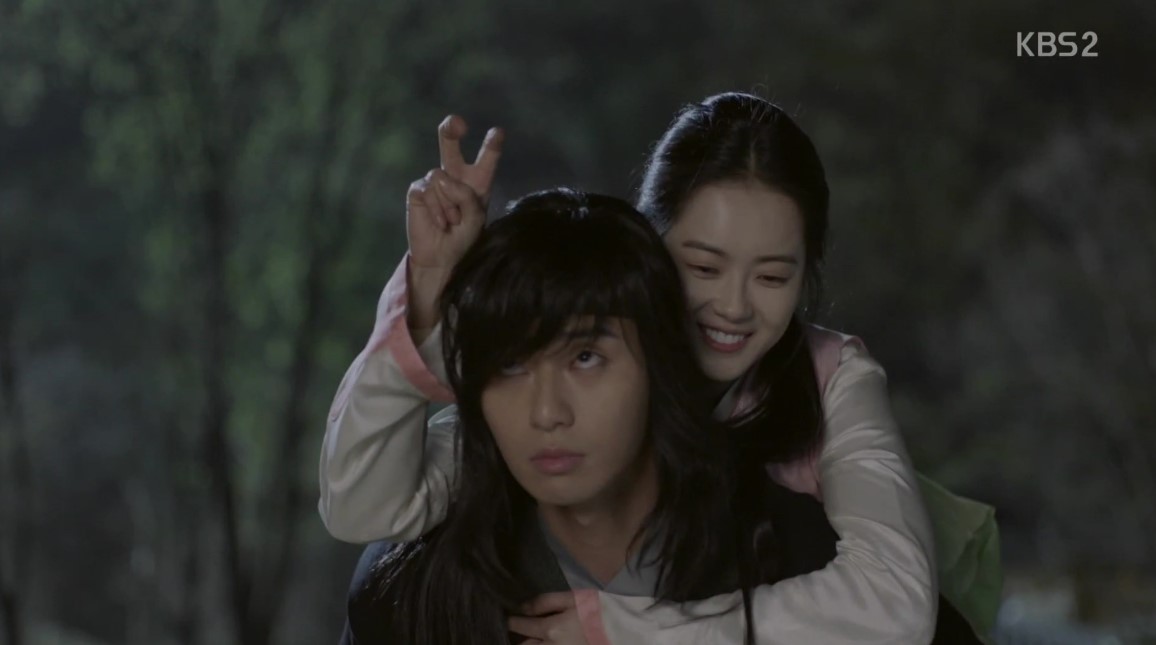 A playful and heartwarming scene from Hwarang, where the female lead gives a peace sign behind the male character’s head as he gives a slightly annoyed but endearing look. The couple shares a light-hearted moment while dressed in traditional hanbok, adding to the charm and chemistry between them.