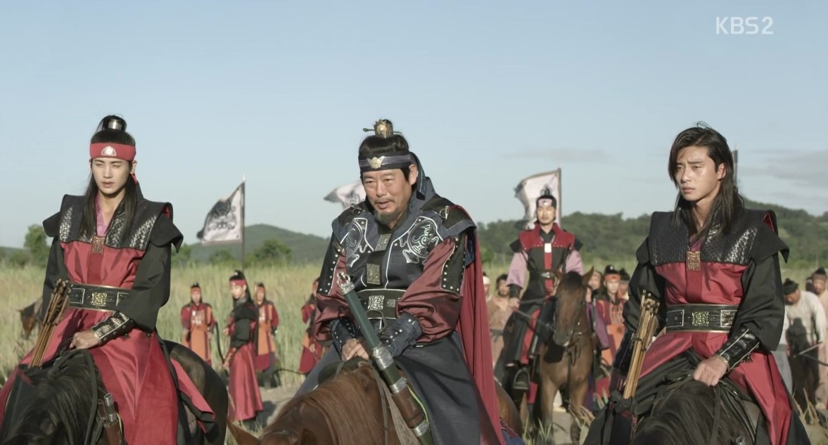 "A powerful scene from Hwarang, showing the main characters and their leader riding horses. The men, dressed in red and black armor, are ready for battle as they lead their army across the open field, showcasing the strength and authority of the Hwarang warriors in this intense moment.