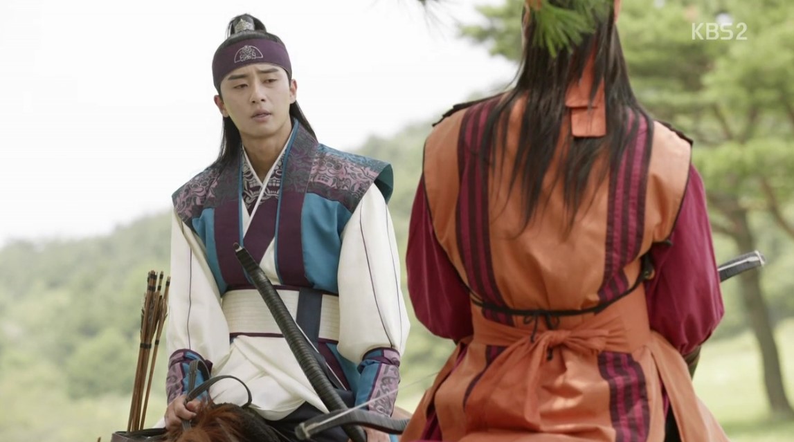 A tense moment from Hwarang, with one of the warriors sitting on horseback while facing another character. The scene conveys a sense of seriousness as they engage in a conversation, with both dressed in traditional attire. The background features natural greenery, setting the mood for the dialogue.