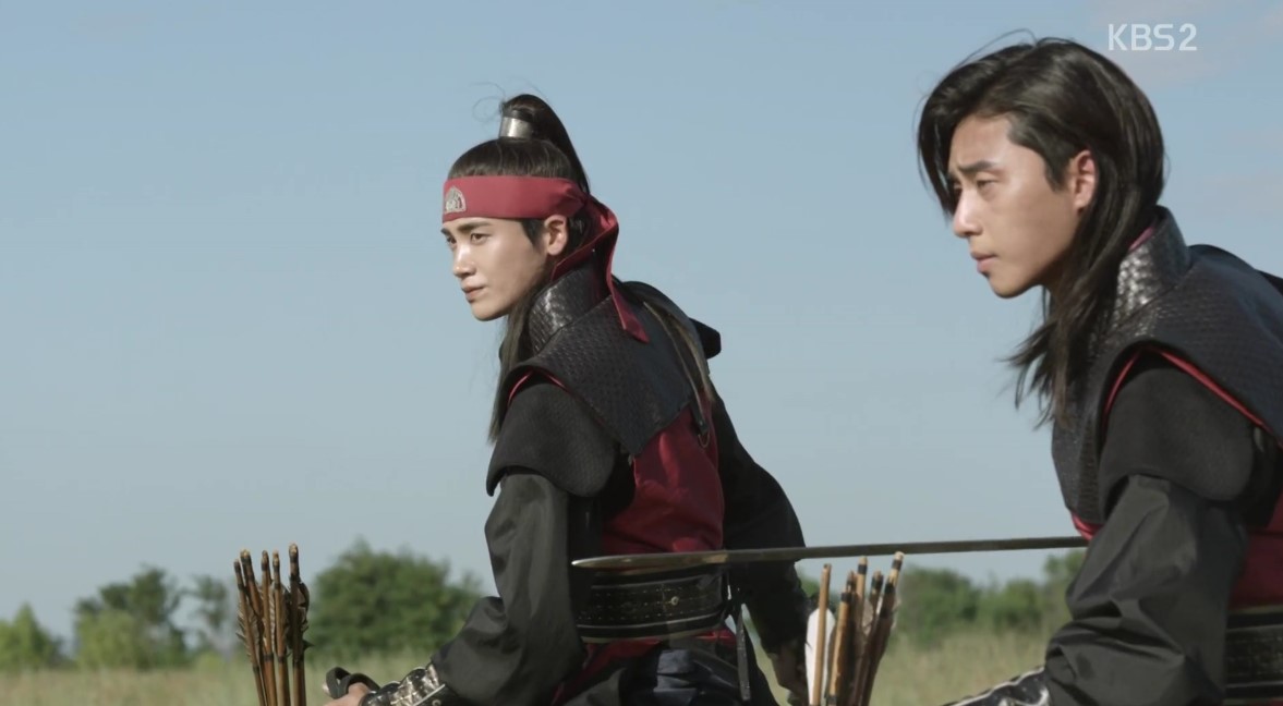 Two warriors from Hwarang on horseback, dressed in red and black armor. They are seen in a focused and determined stance, with bows and arrows at the ready, suggesting an upcoming battle. The bright blue sky and vast open field provide a dramatic backdrop to this intense moment.