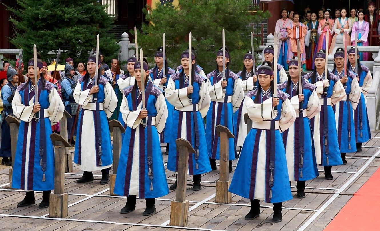 A powerful group of Hwarang warriors lined up in a disciplined formation, dressed in traditional blue and white hanbok. Each warrior holds a sword, standing in perfect alignment, as they participate in a ceremonial or training scene. The vibrant background includes other characters in colorful attire, adding to the intensity and depth of the moment.