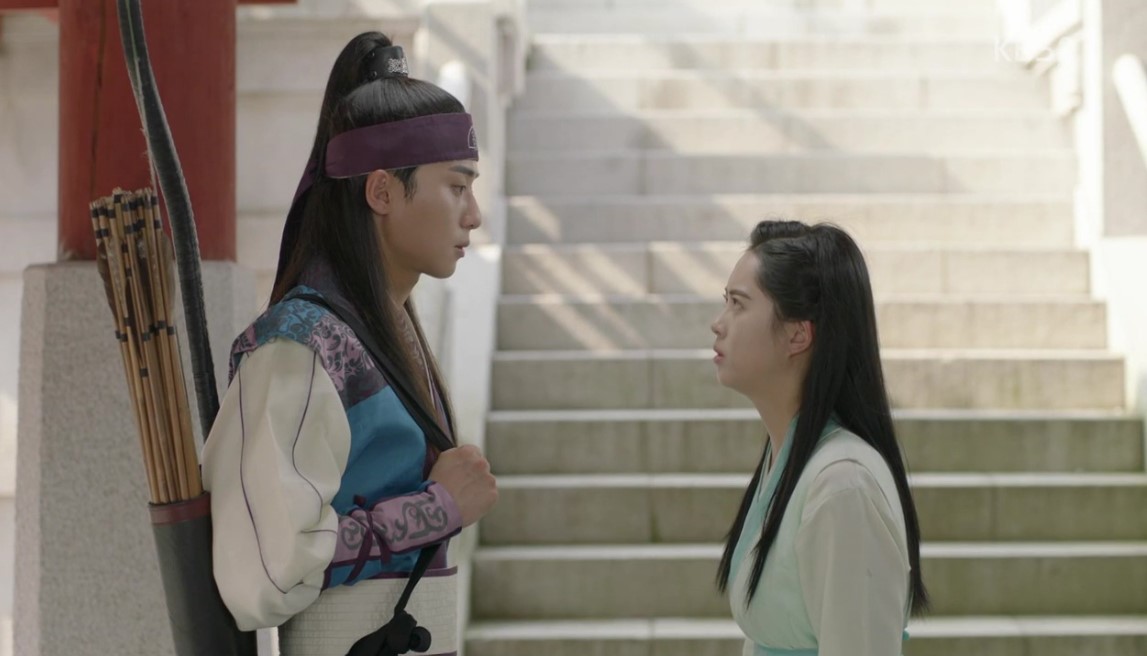 A tense and intimate moment from Hwarang, where the male lead, carrying a quiver of arrows, stands tall in front of the female lead. They lock eyes in a quiet but intense exchange, while the serene backdrop of stairs and architectural details adds to the emotional atmosphere of the scene.