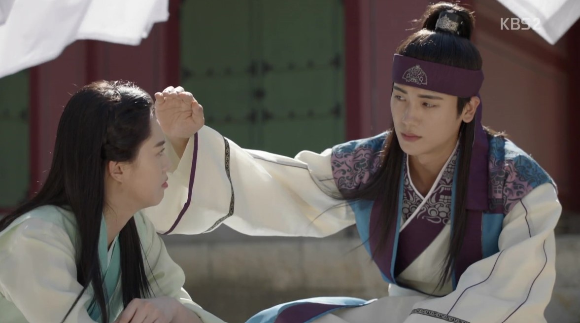 A tender moment from Hwarang, where one of the male leads gently places his hand on the head of the female lead, showing a caring and intimate gesture. Both are dressed in traditional hanbok, and the peaceful setting adds to the calm and affectionate atmosphere of the scene.