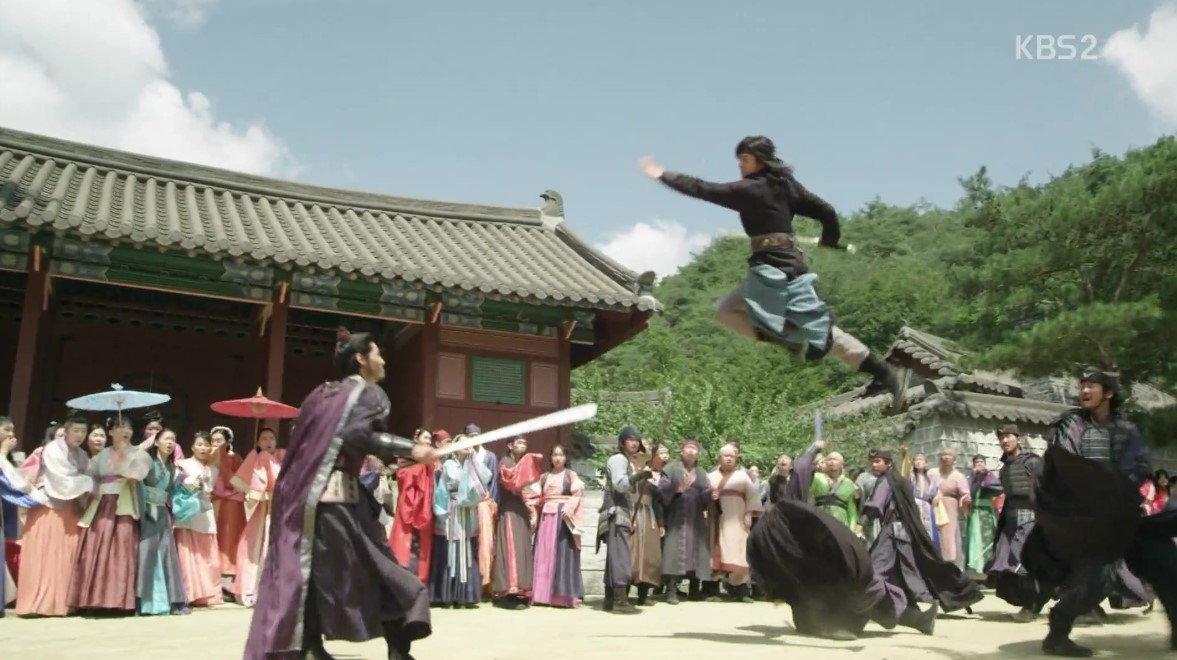 An intense action scene from Hwarang, where two warriors engage in a dramatic sword fight, one leaping high into the air with impressive agility. The battle takes place in front of a crowd, with spectators in colorful attire watching the fierce demonstration of skill and strength.