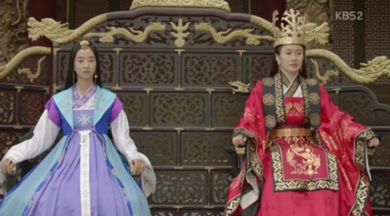 A royal scene from Hwarang, showing two powerful women seated on thrones. One is dressed in a regal red gown with a crown, representing authority, while the other wears a more modest yet elegant purple hanbok, symbolizing a quieter strength. The intricate throne and the grandeur of the setting emphasize the importance of their roles in the series.