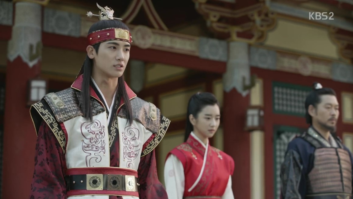 A powerful scene from Hwarang, with the main character dressed in elaborate armor standing confidently. He is flanked by a strong female character in a red traditional gown, and another man in regal attire. The background highlights the grandeur of the palace, enhancing the sense of leadership and authority.