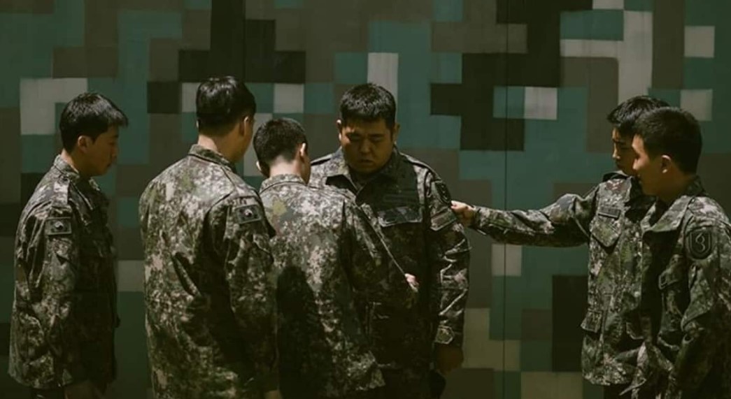 Scene from DP Season 1 showing a group of soldiers in military uniform, with one soldier being bullied by his comrades in front of a camouflaged wall. This tense moment reflects the harsh realities and emotional struggles explored in DP Season 1, capturing the bullying and pressure within the military context, as discussed in the article.