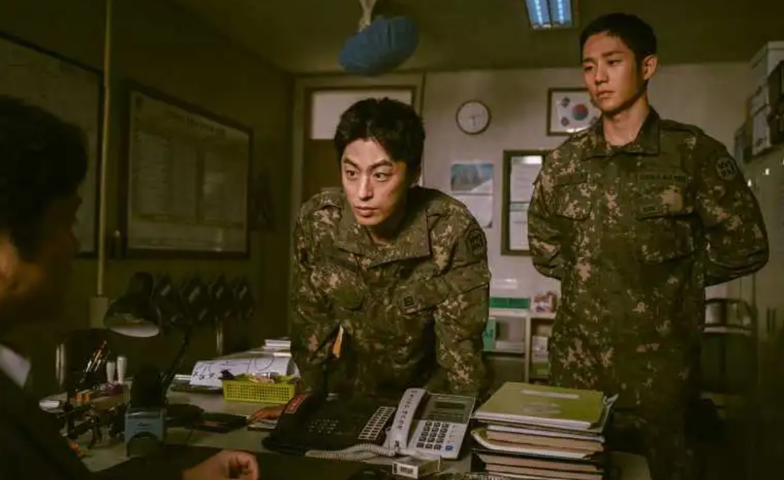 Scene from DP Season 1, showing two soldiers in military uniform, standing in a tense moment inside an office. One soldier is leaning forward with a serious expression while the other stands silently, reflecting the intense power dynamics and conflict depicted in the drama. This moment encapsulates the harsh military atmosphere explored in the article.