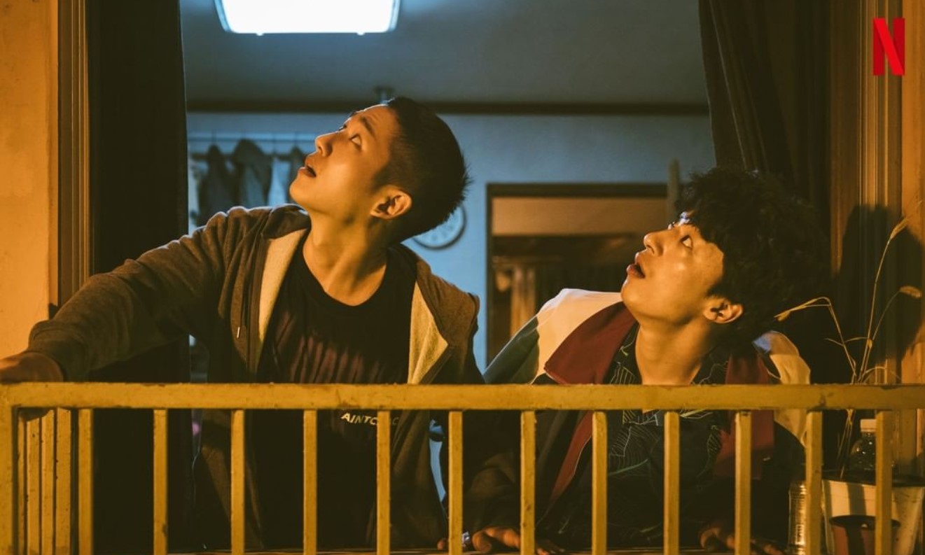 DP Season 1 scene showing two characters standing on a balcony, looking up at something in the night sky with expressions of concern or curiosity. The tense atmosphere highlights the dramatic nature of the show, reflecting the suspense and emotional struggles.