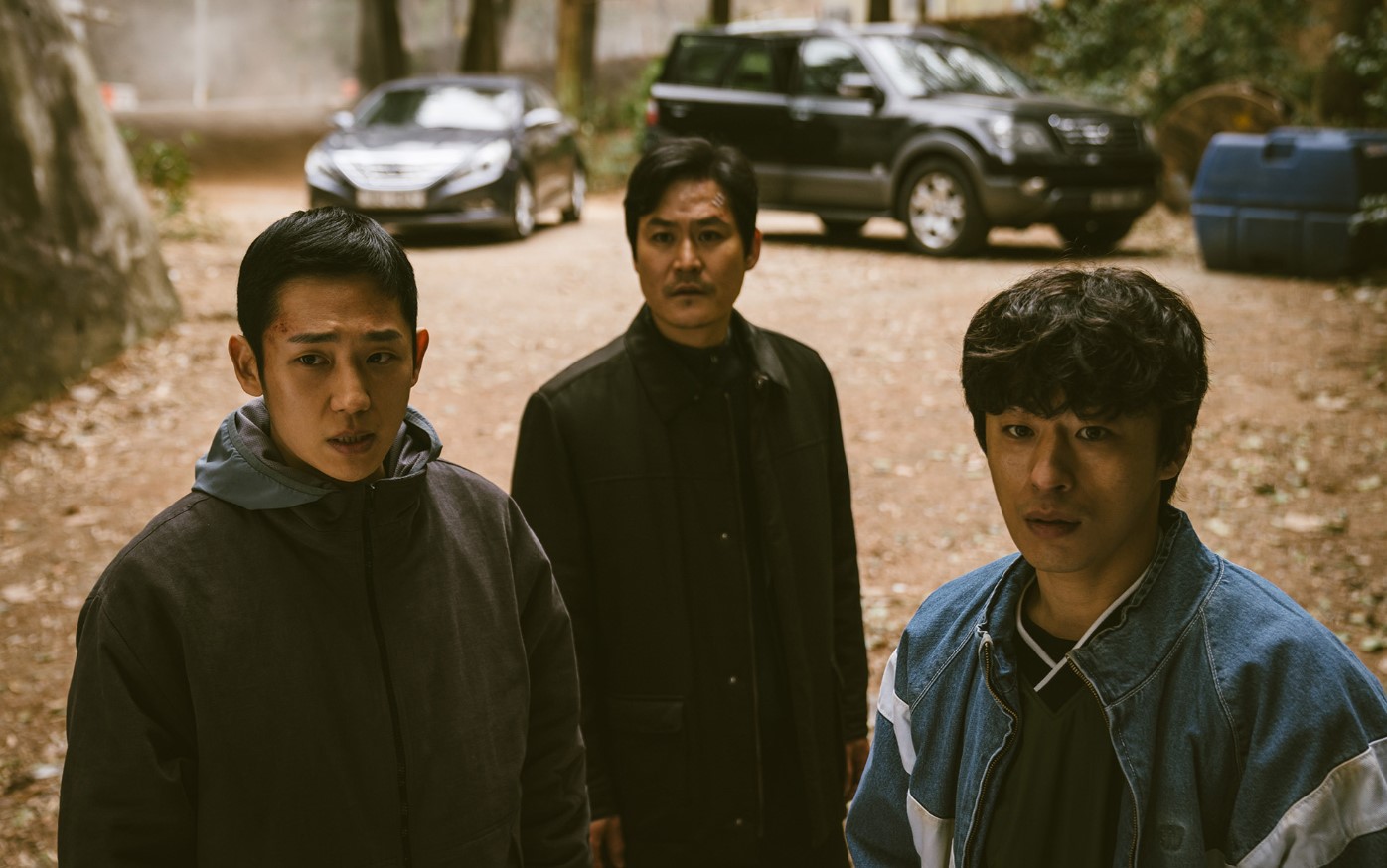DP Season 1 scene showing three characters in a tense moment, standing in a wooded area with two cars parked in the background. The characters' expressions reflect the gravity of the situation, with one visibly bruised, embodying the intense drama and conflict explored in DP Season 1. This powerful image is in line with the themes discussed.