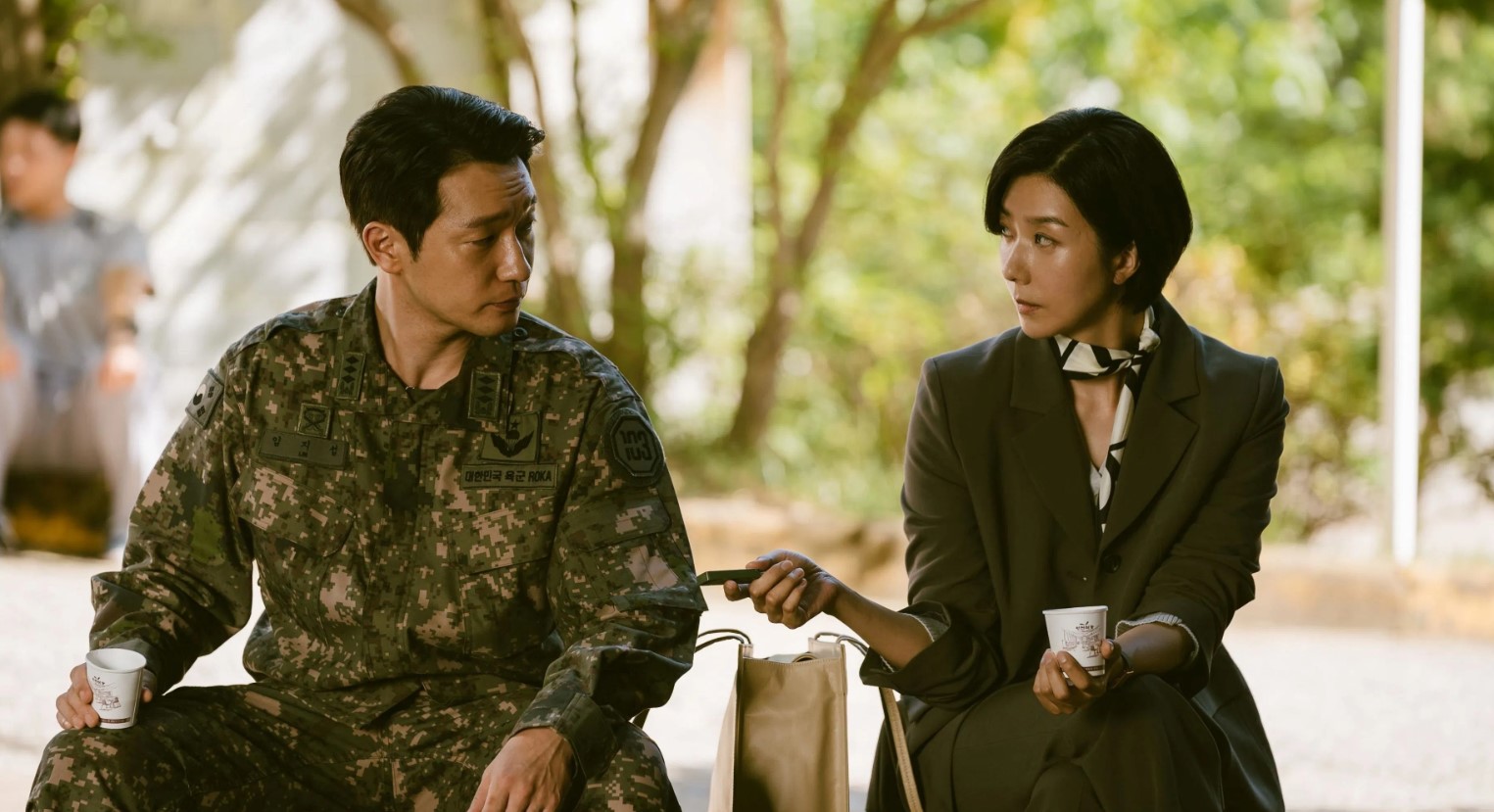 Scene from D.P. season 2 showing a soldier in uniform sitting with a woman in formal attire, having an intense conversation in a peaceful outdoor setting. The woman is handing the soldier her phone, with both characters conveying emotions of curiosity and tension. Background features blurred greenery, creating a calm yet dramatic atmosphere.