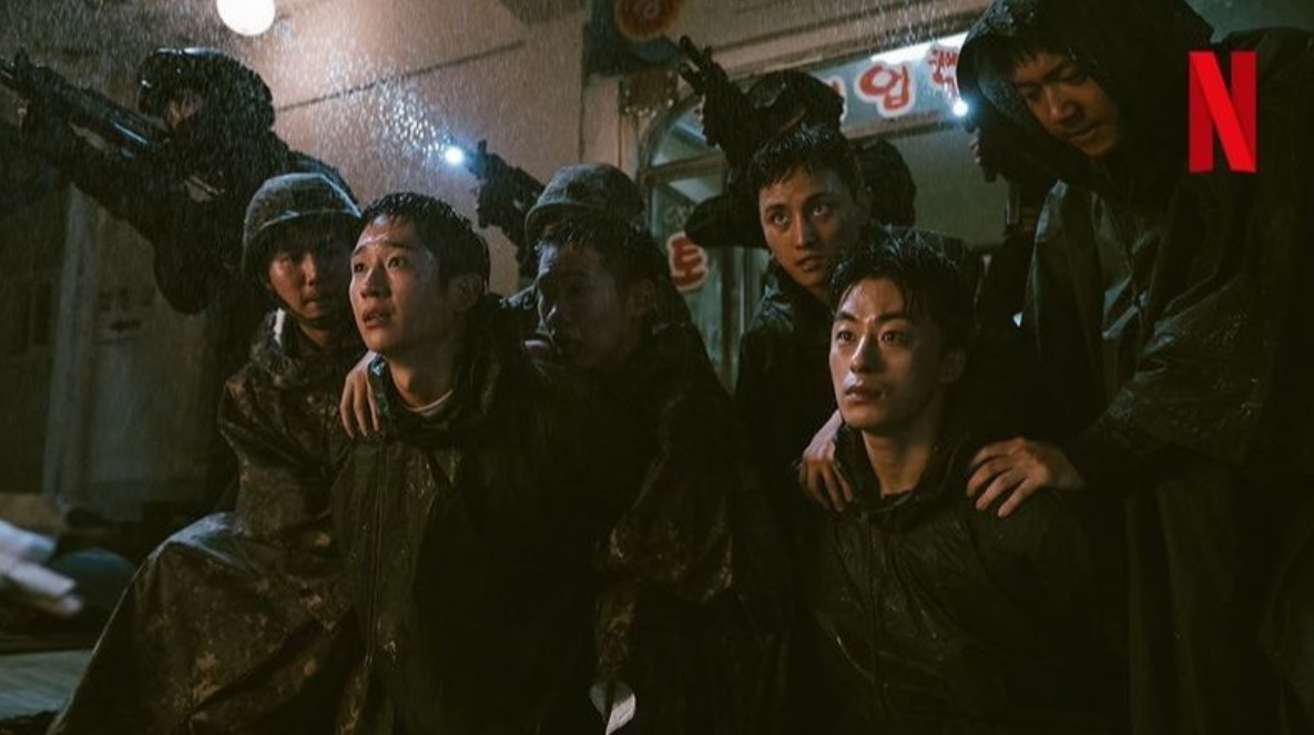 Scene from 'D.P. Season 2' showing a group of soldiers in rain-soaked gear, huddled together in a tense moment. One soldier sits in the foreground with a distressed expression, while others look around, seemingly under threat. Armed soldiers are visible in the background, creating a dark, suspenseful atmosphere. The Netflix logo appears in the top right corner