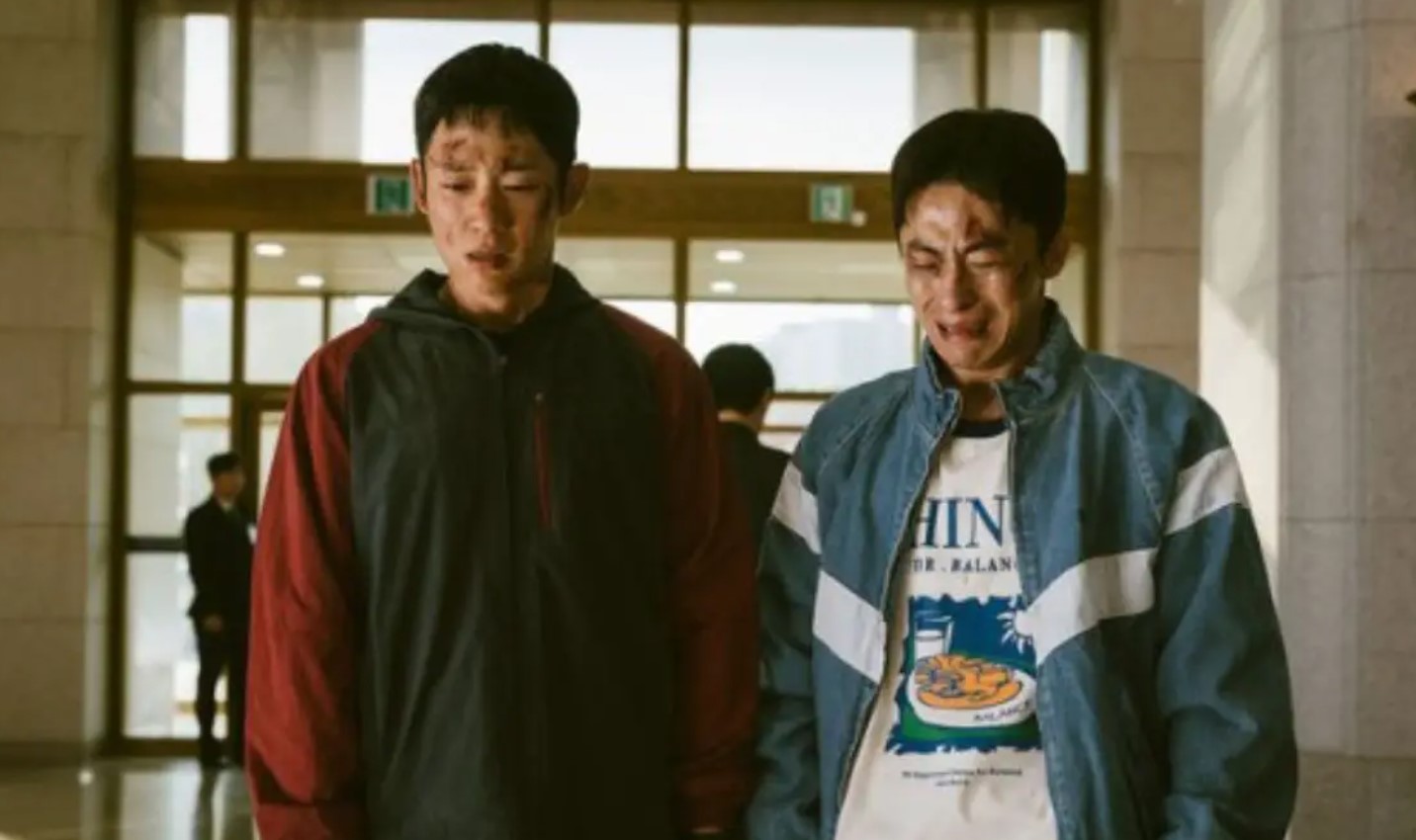 Scene where two characters standing with bruises and dirt on their faces. The expressions of the characters show distress and pain, highlighting the intense moment. They are in a large, well-lit building, possibly after a confrontation, with figures in the background.