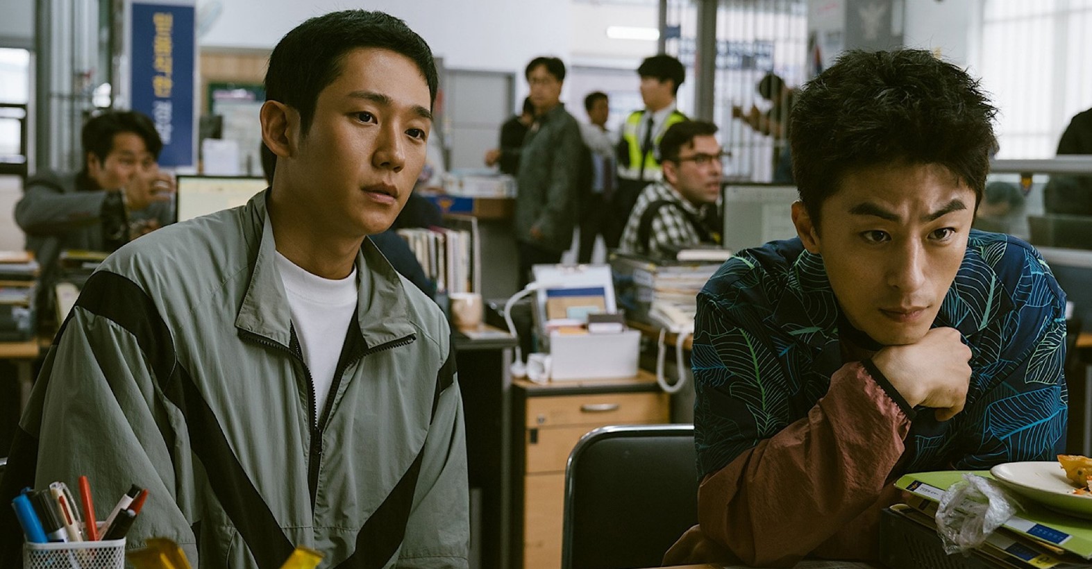 Two characters sitting in a busy office, both deep in thought. One character looks at the camera while the other has his hand on his chin. In the background, office workers are seen engaged in their tasks. The setting creates a tense yet everyday atmosphere.
