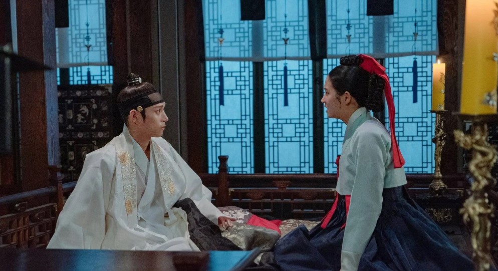 Intimate scene from The Forbidden Marriage, showing the male and female leads sitting across from each other, deep in conversation, with traditional Korean furnishings and soft lighting creating a serene atmosphere.