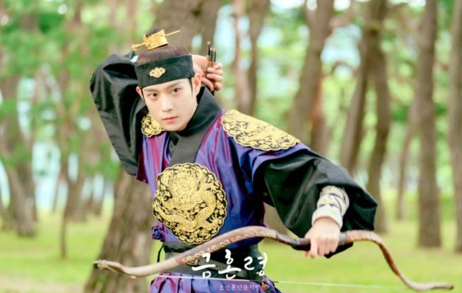 Action-packed scene from The Forbidden Marriage, showing the male lead in royal archery attire, aiming his bow in a forested area, highlighting his determined expression and skillful stance.