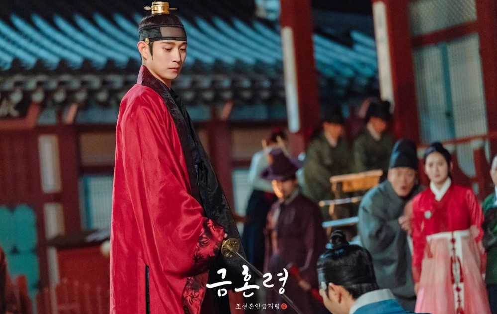 Intense courtroom scene from The Forbidden Marriage showing the male lead in a regal red robe, standing tall and gazing downward as others watch, set against a traditional Korean palace backdrop.