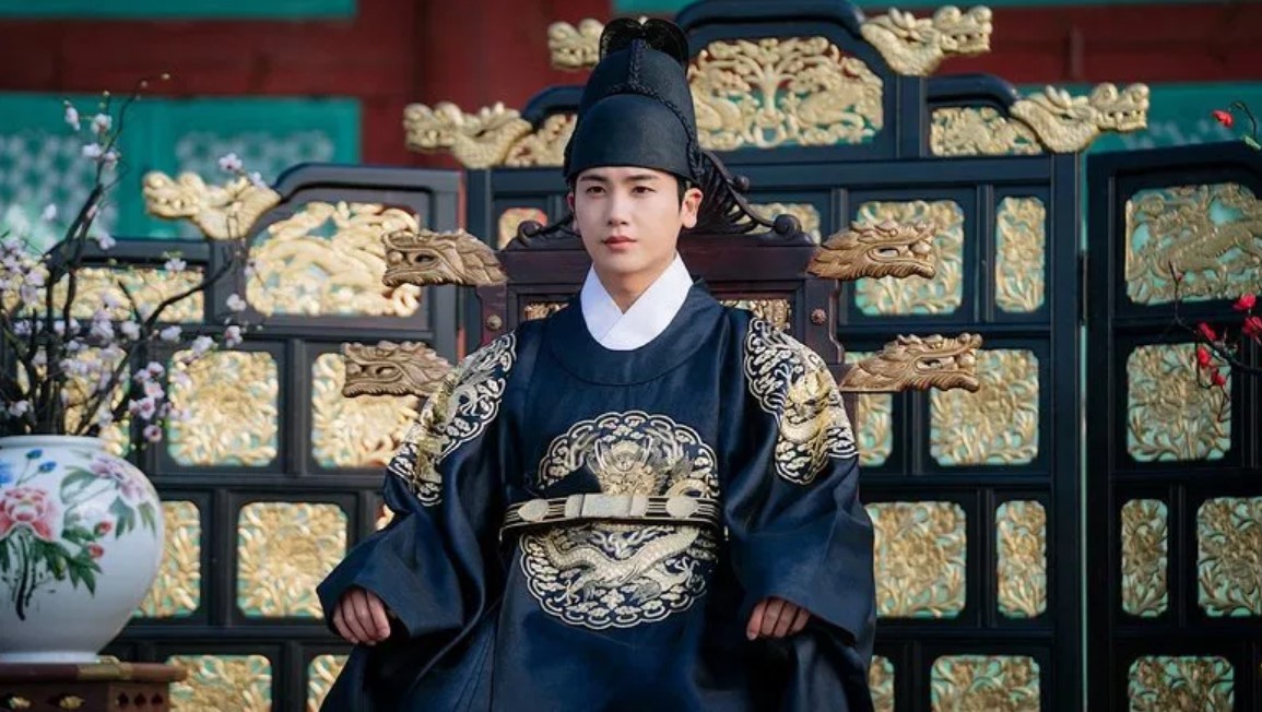  The male lead from Our Blooming Youth sitting confidently on a throne, dressed in an ornate royal robe, surrounded by elegant decorations.