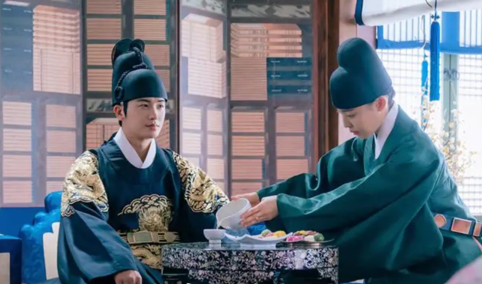 Two characters from Our Blooming Youth are seated at a table, one in a royal robe while the other is serving food. The setting is elegant, with traditional decor and colorful treats displayed, highlighting a moment of interaction between them. 