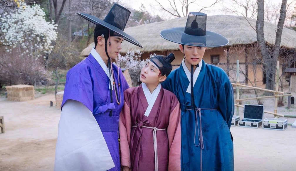 Three main characters from Our Blooming Youth standing together outdoors, with the female lead in between the two male leads. The setting features traditional hanbok attire, with the backdrop of a serene, natural environment.