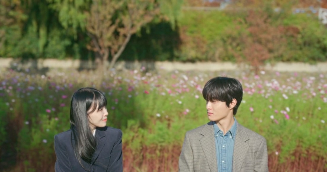  A peaceful scene from The Delivery Man, with the two main characters sitting on a bench in a field of flowers. They gaze at each other, sharing a moment of connection amidst the serene natural surroundings.