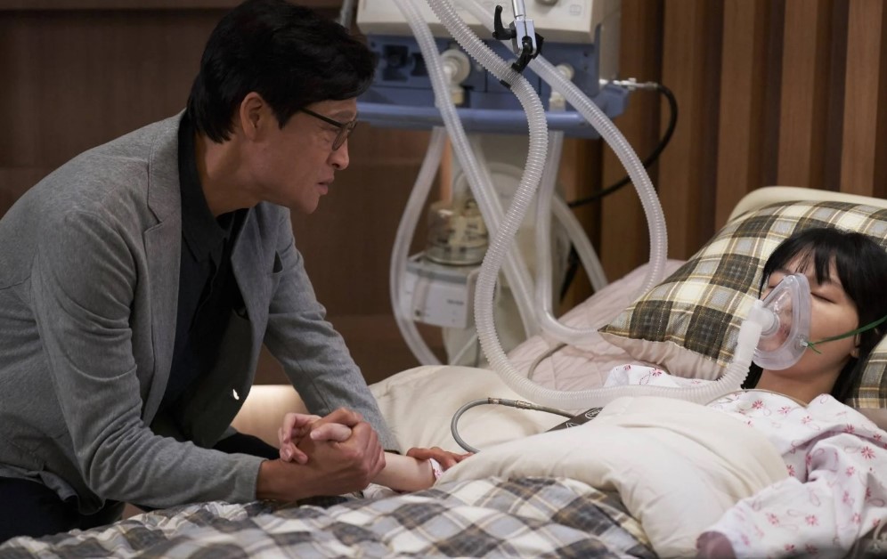 A touching scene from The Delivery Man, showing a man holding the hand of a woman lying unconscious in a hospital bed, connected to life support. The man looks at her with concern, highlighting an emotional moment in the drama