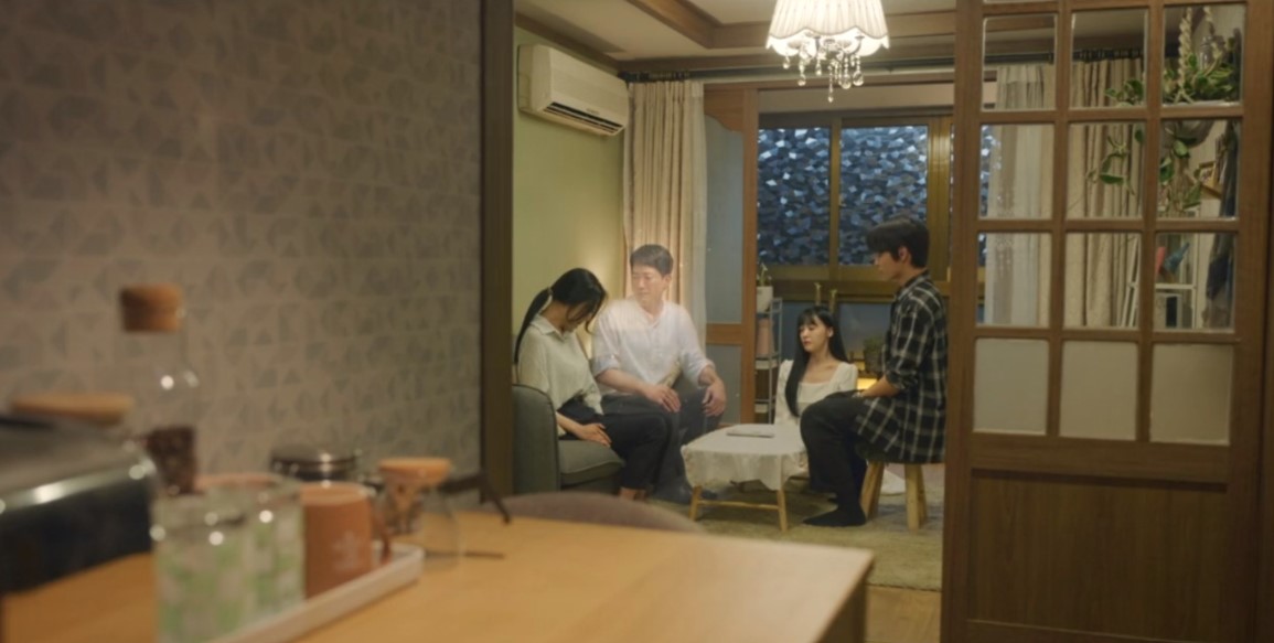 A cozy domestic scene from The Delivery Man, showing four characters in a living room. Two are sitting on a sofa, while the other two are seated at a small table, all looking slightly pensive in the warm, intimate setting.