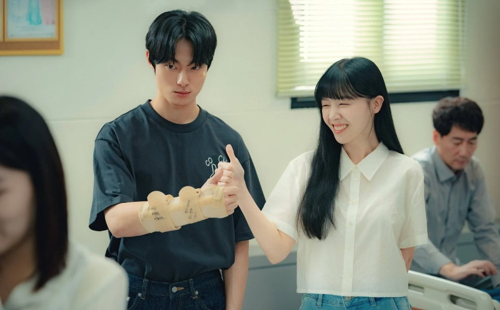 A playful moment from The Delivery Man, showing the two main characters in a lighthearted interaction. The male character, wearing a t-shirt and a bandaged hand, looks serious, while the female character, smiling and winking, gives him a thumbs-up. They stand together in a casual setting with other people in the background.
