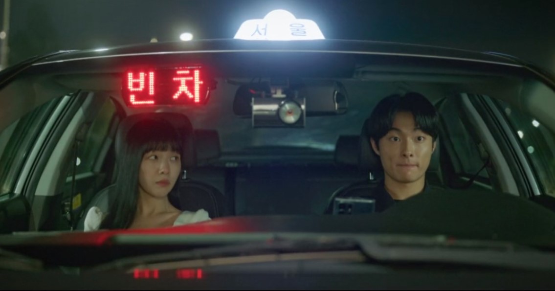 A scene from The Delivery Man showing the two main characters sitting in a taxi. The woman looks out the window with a slightly serious expression, while the man glances at her, appearing a bit nervous. The taxi's "빈 차" (empty) sign is illuminated in the background.