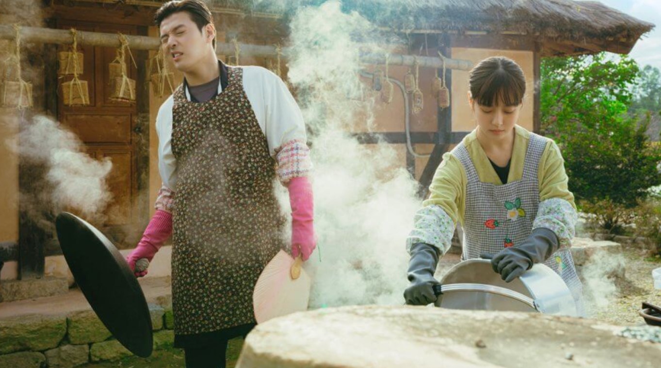 A humorous and charming scene from Tastefully Yours, showing the main characters cooking together in a traditional setting, with steam rising from their dishes as they wear matching aprons and gloves.