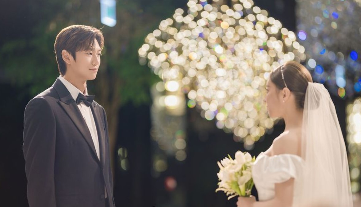 A magical and romantic moment from a Korean drama, showing a groom in a tuxedo standing in front of his bride in a wedding dress, both surrounded by a beautiful background of sparkling lights.