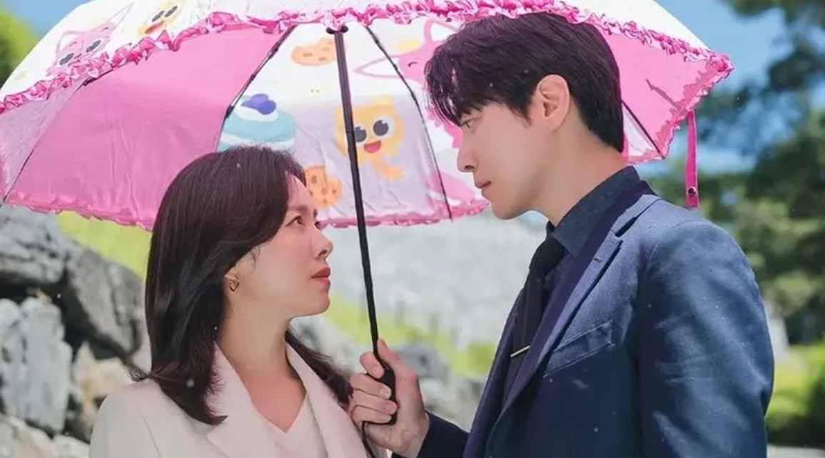A sweet and intimate moment from a Korean drama, showing the two main characters sharing a moment under a playful, pink umbrella adorned with cartoon designs, as they gaze into each other's eyes.