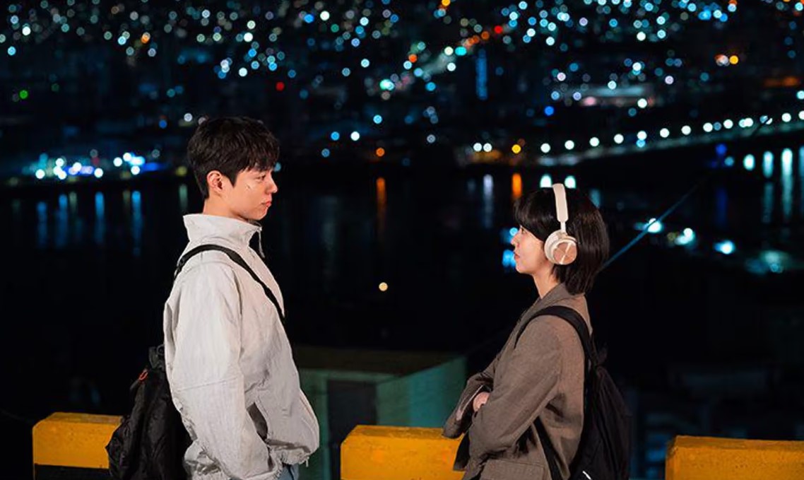 A romantic scene from Good Boy, featuring two characters standing on a bridge at night, with the city lights sparkling in the background. The man looks at the woman, who is wearing headphones and facing him.