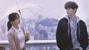 Romantic scene from the Korean drama Just Between Lovers, showcasing a couple in a heartfelt moment. A woman holding an umbrella gazes lovingly at the man, who looks away thoughtfully, against a soft, snowy cityscape backdrop. One of the most underrated Korean dramas that captures deep emotions and complex relationships.