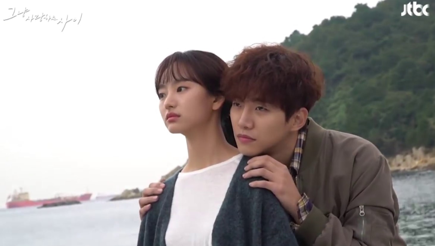 Emotional scene from the Korean drama Just Between Lovers, featuring the main couple sharing a quiet, intimate moment by the sea. The woman stands in front, while the man gently wraps his arms around her, displaying their deep connection. One of the most underrated Korean dramas that beautifully portrays love and healing.