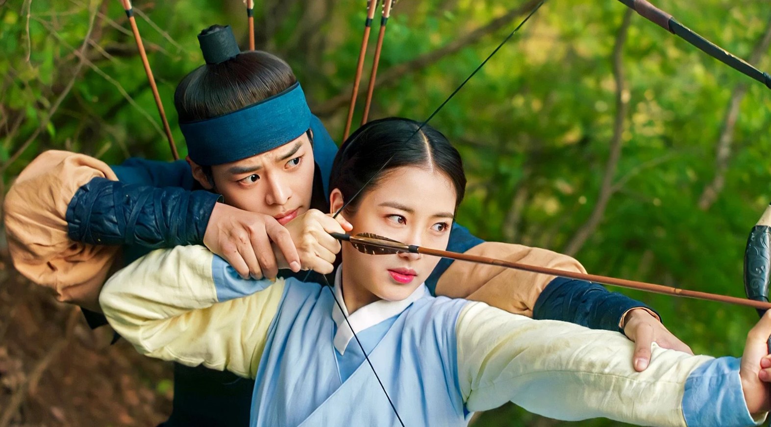 A close-up action scene from "My Secret Romantic Guesthouse," showing a young woman aiming a bow and arrow with intense focus, while a young man stands close behind her, guiding her shot. The background is filled with lush green foliage, adding to the dramatic moment.