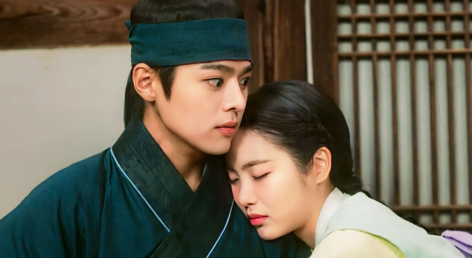 An emotional scene from "My Secret Romantic Guesthouse," showing a young woman resting her head on a young man's chest with her eyes closed, while he looks ahead with a serious and protective expression. The moment captures a tender connection between the two characters.