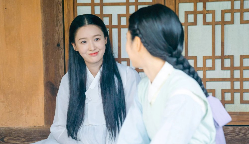 A scene from "My Secret Romantic Guesthouse," showing two women in traditional attire seated and talking. One woman with long black hair smiles warmly at the other, creating a light and friendly moment. Great image for "My Secret Romantic Guesthouse full review.