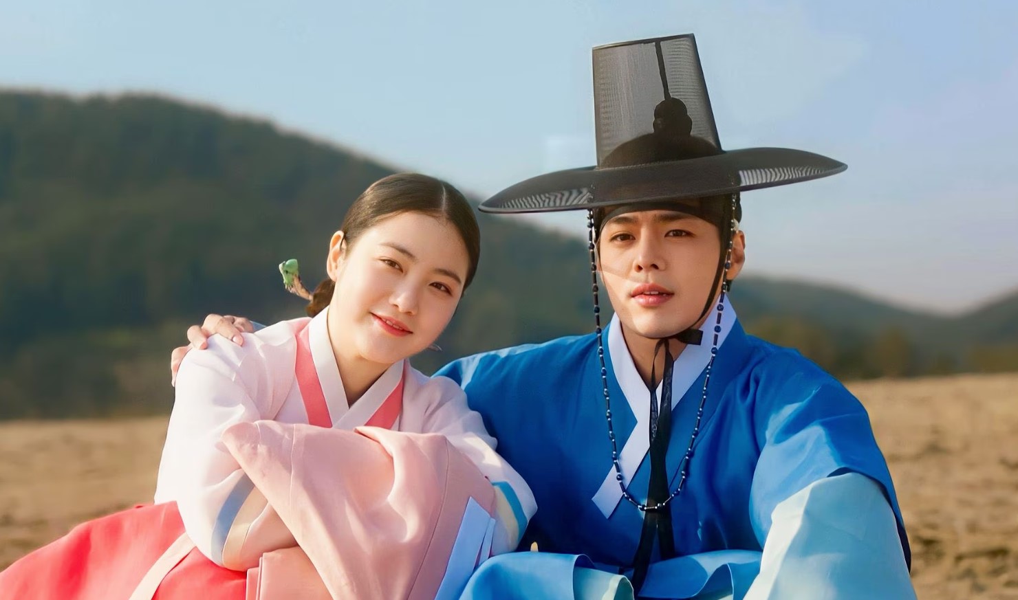 A bright and cheerful scene from "My Secret Romantic Guesthouse," featuring a young woman in a pink hanbok and a young man in a blue hanbok with a traditional hat. They sit closely together outdoors, smiling with a scenic mountain landscape in the background.