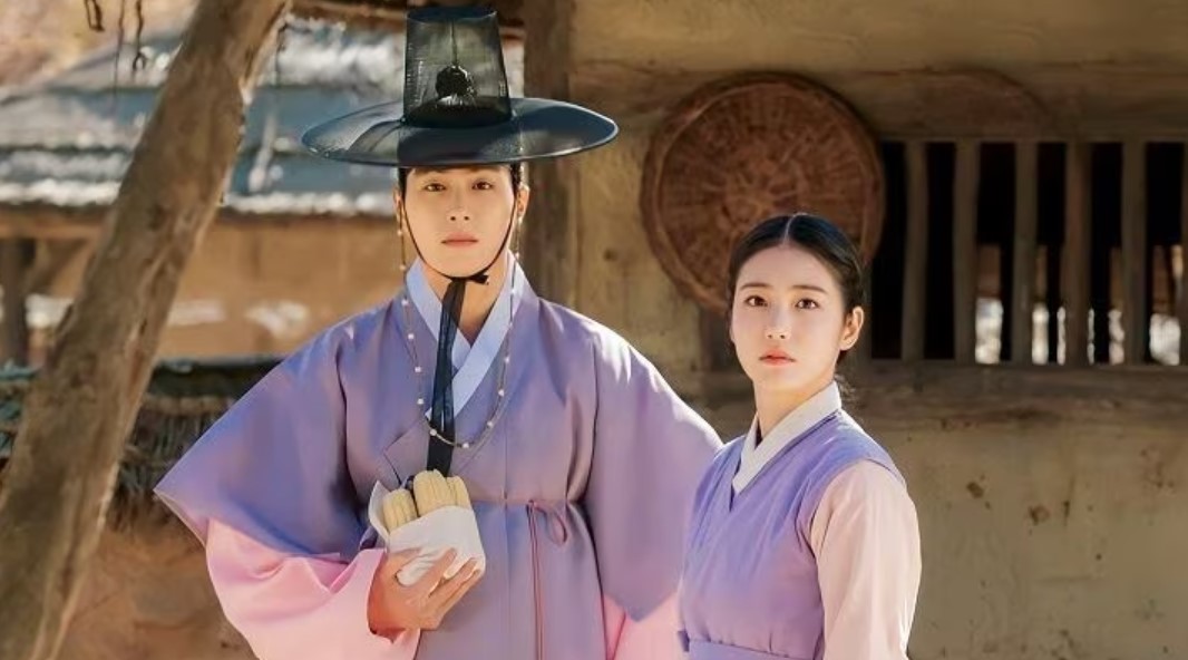 A scene from "My Secret Romantic Guesthouse," showing a young man in a light purple hanbok and traditional hat holding a bundle of corn, standing beside a young woman in a matching purple outfit. They both look ahead with calm, thoughtful expressions in front of a rustic village backdrop.