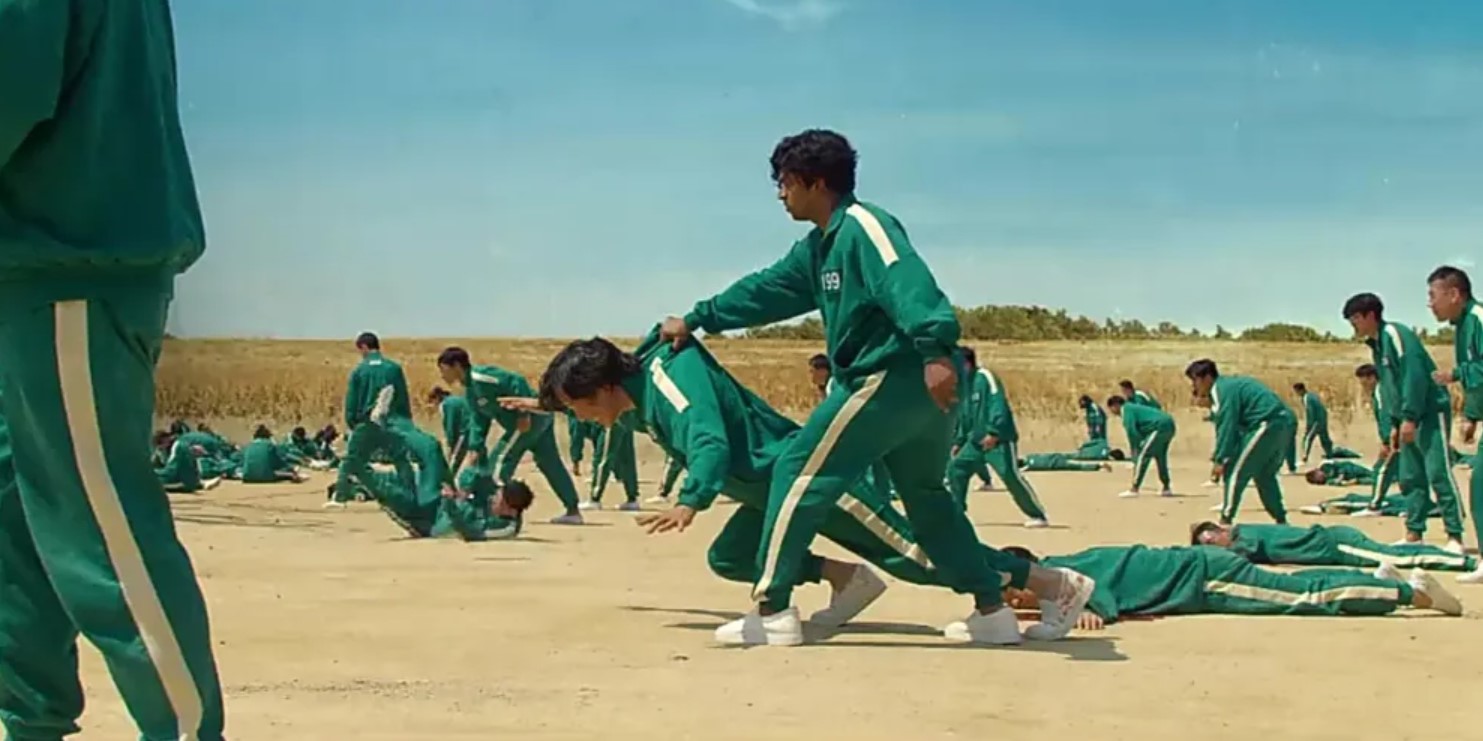 Contestants in Squid Game Season 1, dressed in green tracksuits, participate in a tense moment of the deadly game. One player is helping another while others are scattered on the ground, capturing the high stakes of the competition. This moment highlights the desperation and tension of the Squid Game challenge, perfect for a Squid Game season 1 full review.