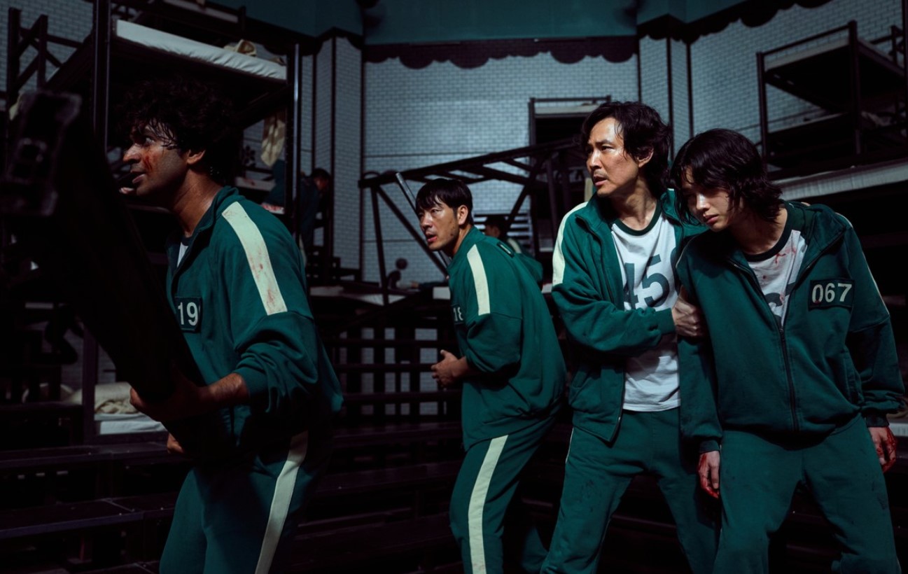 Contestants in Squid Game Season 1, dressed in green tracksuits, including numbers 19, 45, and 067, are seen in a tense moment as they prepare for action. The group, bloodied and weary, stands in front of a complex metal structure, showcasing the danger and high-stakes nature of the deadly game.