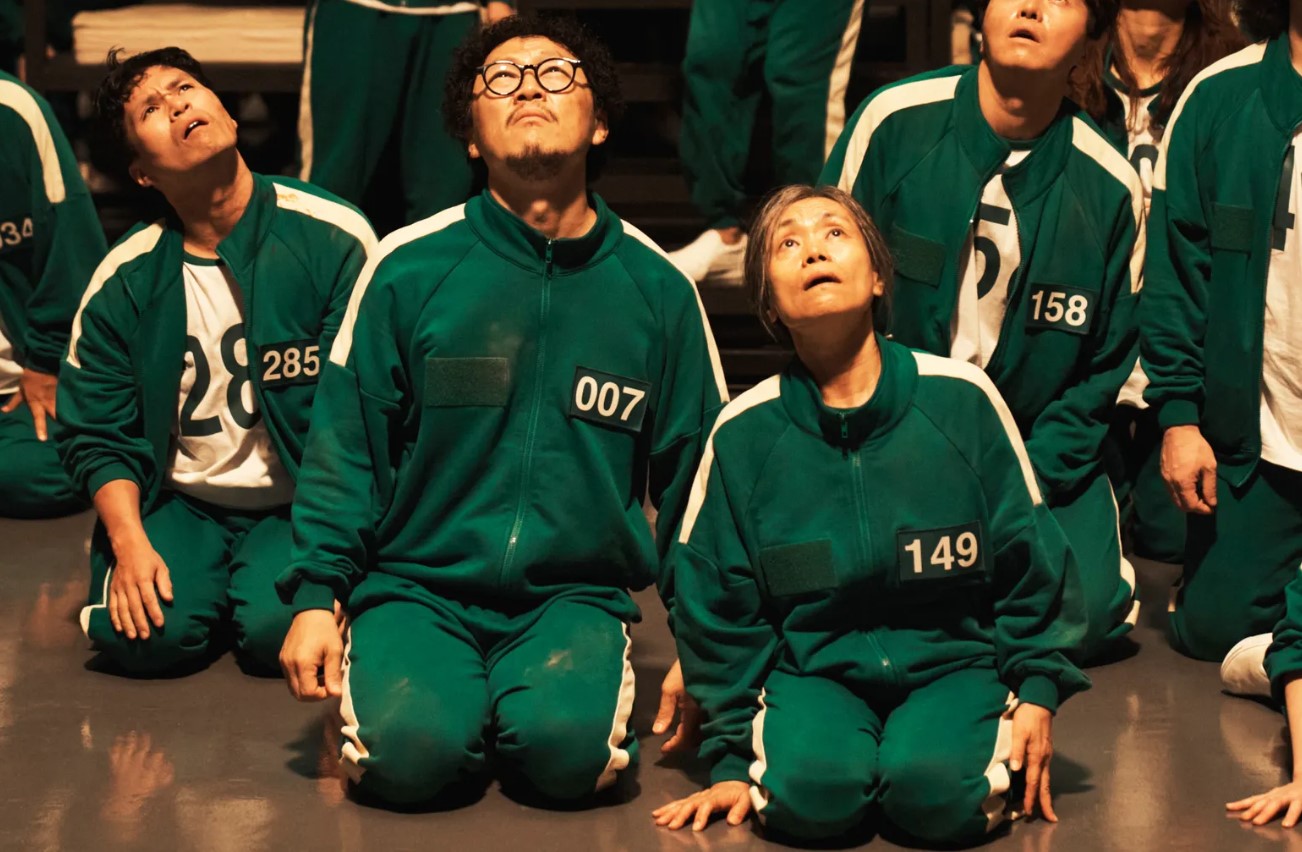 Dramatic moment in Squid Game Season 2 showing contestants kneeling and gazing upwards, filled with anticipation and fear as they face an uncertain fate.