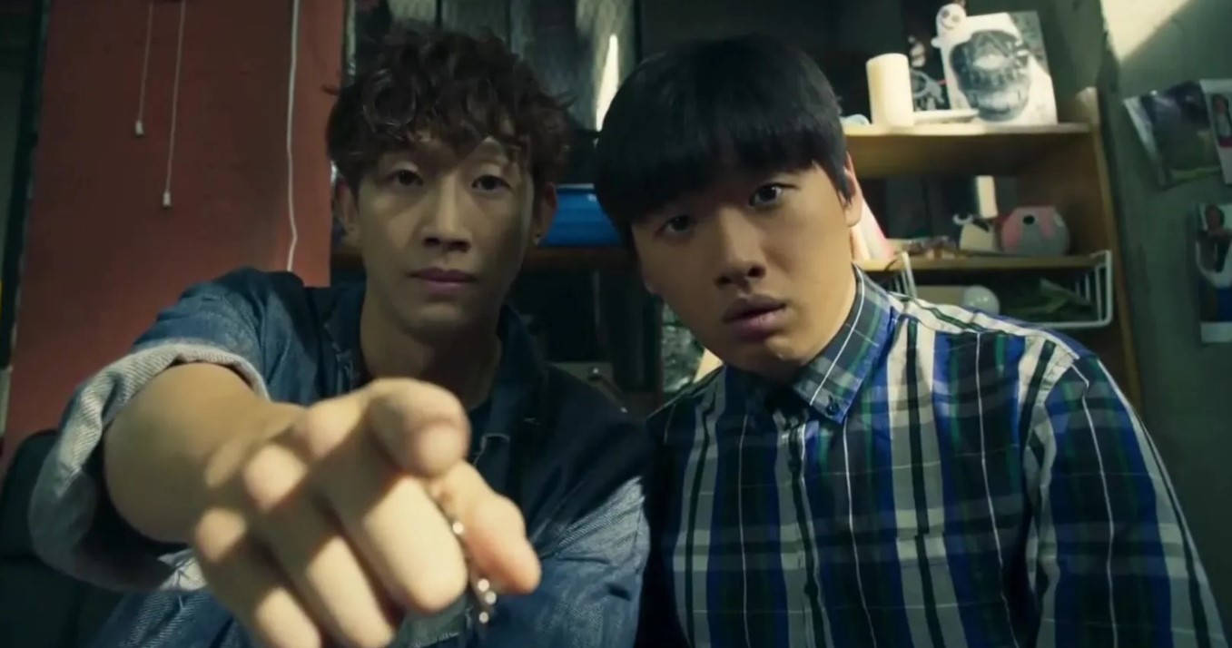 Scene from the Korean drama Let’s Fight the Ghost, featuring two male characters sitting together in a room. One is pointing towards the camera with a serious expression, while the other looks surprised or concerned. Both are dressed casually, with the background showing various objects and cluttered shelves. Let’s Fight the Ghost full review.