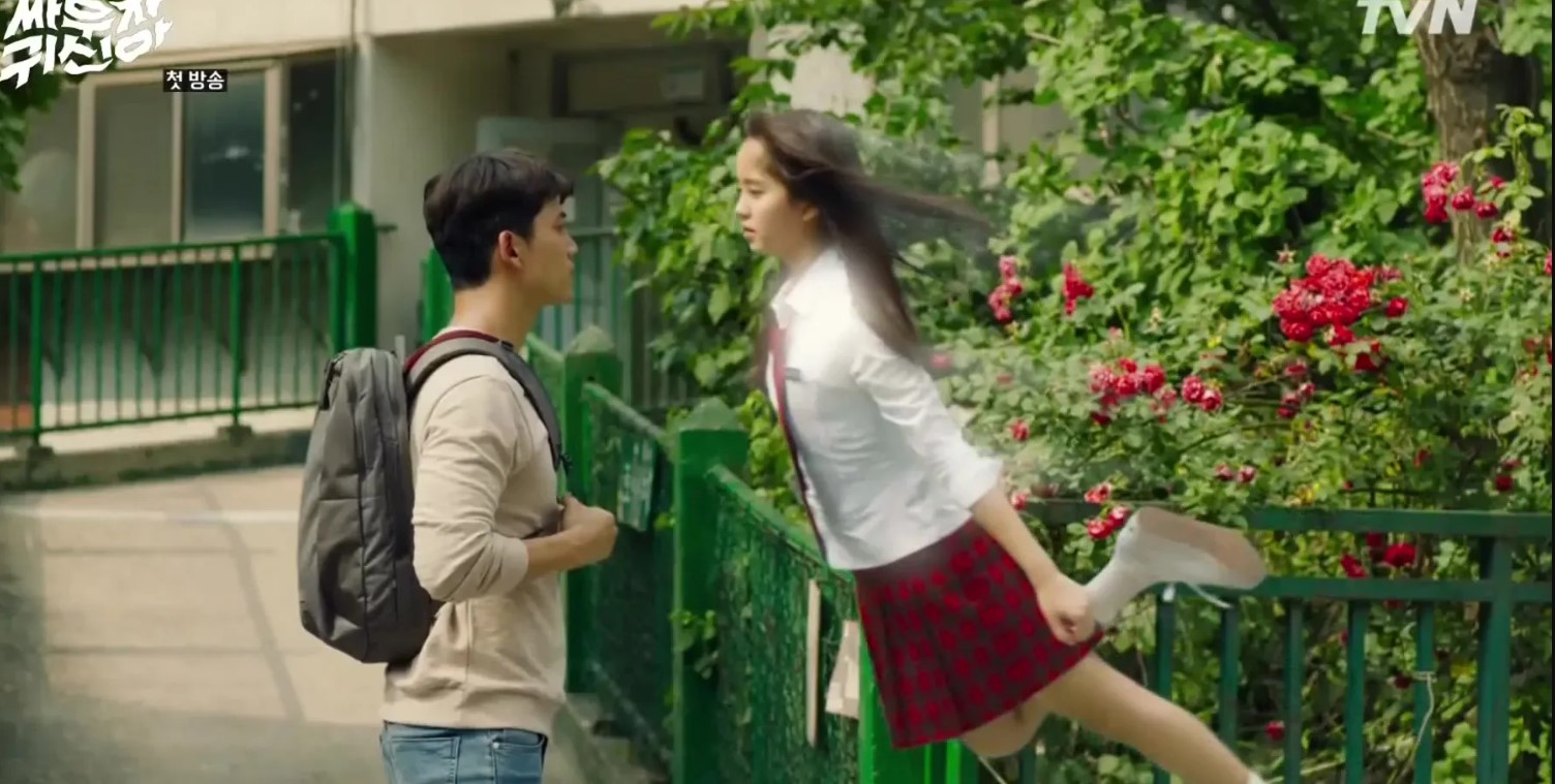Scene from the Korean drama Let’s Fight the Ghost, showing a ghostly female character in a school uniform floating in mid-air near a male character, who appears surprised. They stand outside in front of a green fence, with lush greenery and red flowers in the background. The female character’s movement is enhanced with a blur effect, emphasizing her ethereal nature.