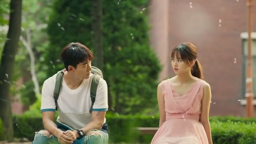 A scene from the Korean drama Let’s Fight the Ghost, showing the two main characters sitting on a park bench. The male character is looking at the female character, who is facing forward with a slightly indifferent expression. The background shows trees and greenery, with soft, floating petals or pollen in the air, adding a serene atmosphere to the moment. 